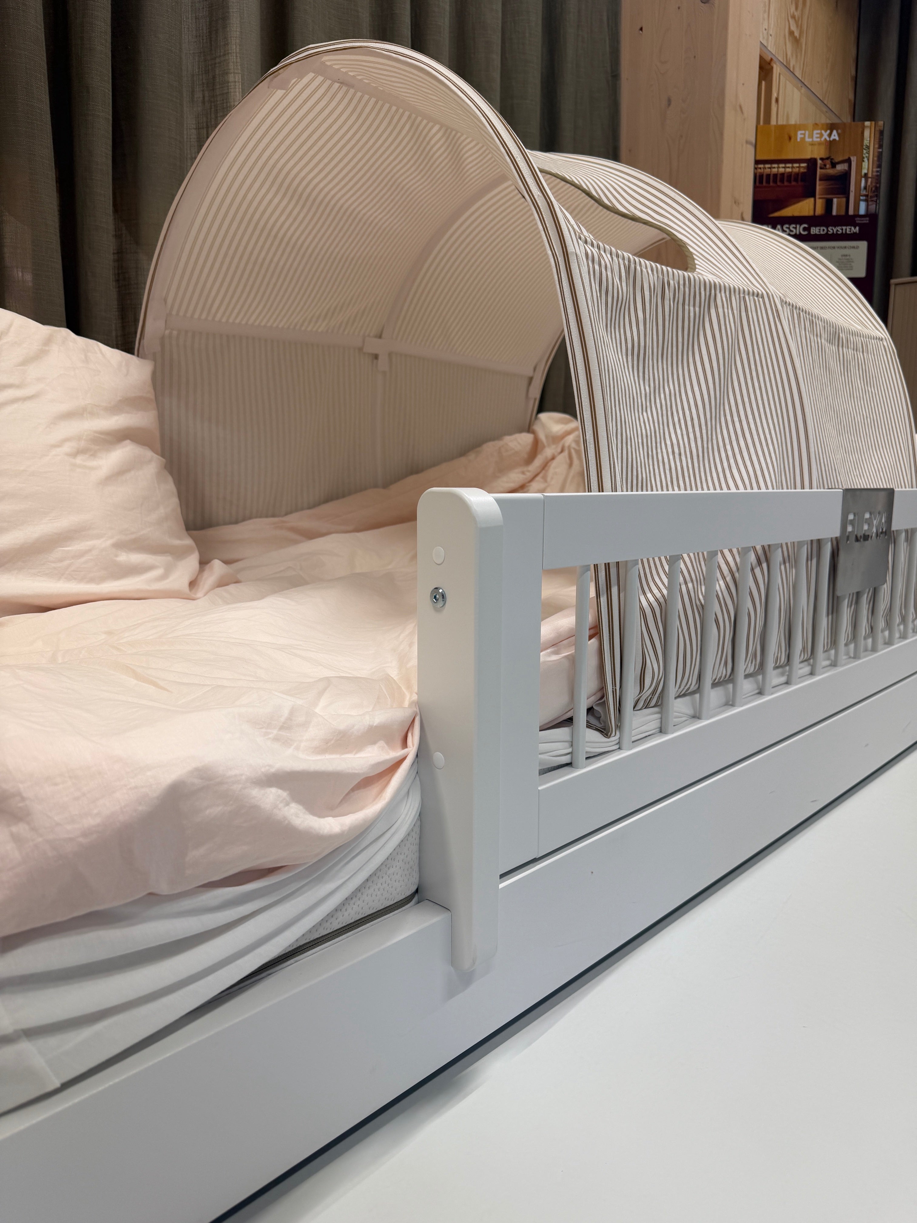 FLEXA white children’s bed with safety rail, striped canopy, pink bedding and books on shelf in a child’s room.