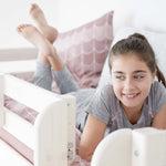 Girl smiling on FLEXA High Bed 184 cm, white frame, pink bedding; bright room with desk, sofa, and safety rail visible.