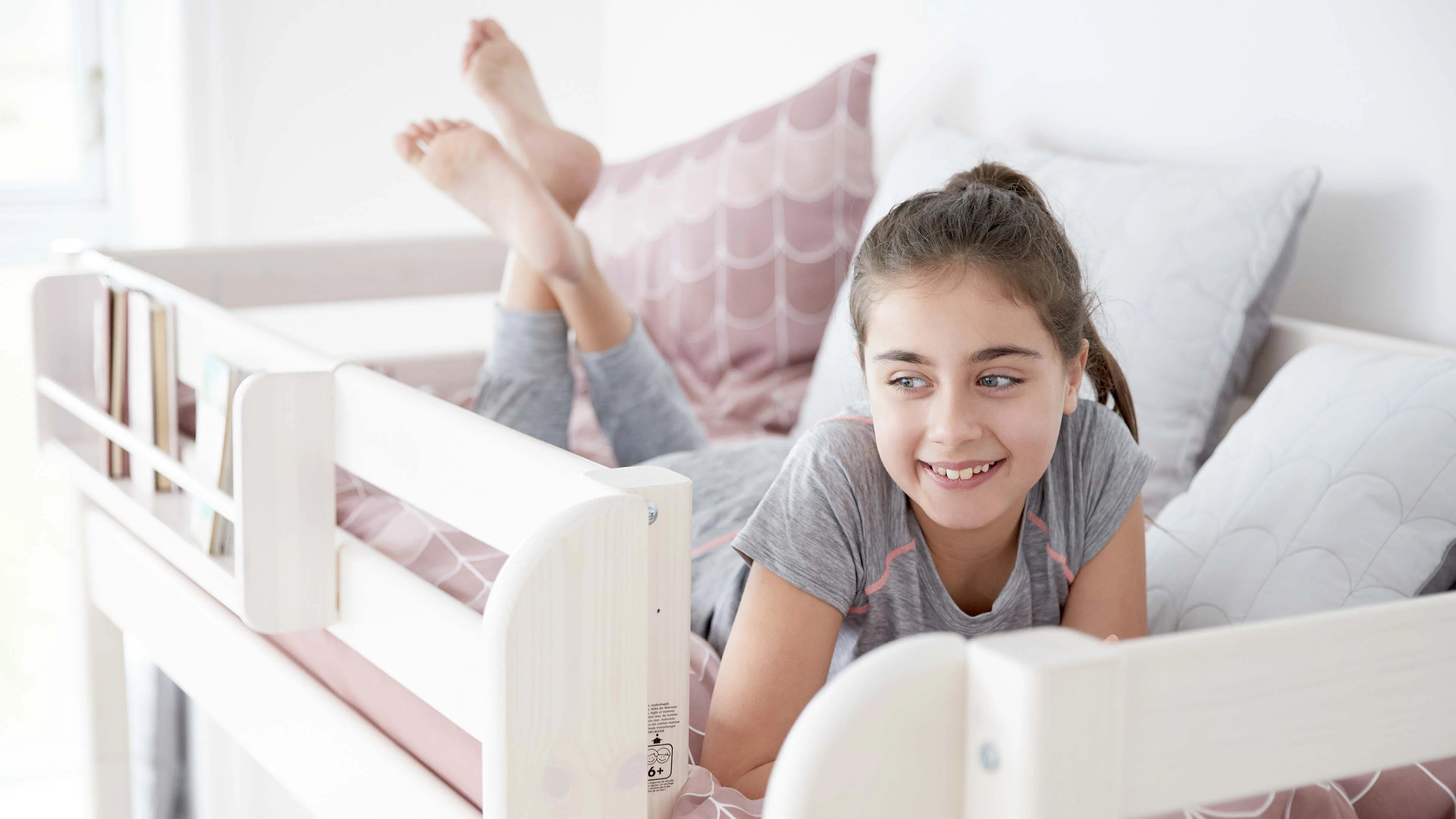 Girl smiling on FLEXA High Bed 184 cm, white frame, pink bedding; bright room with desk, sofa, and safety rail visible.